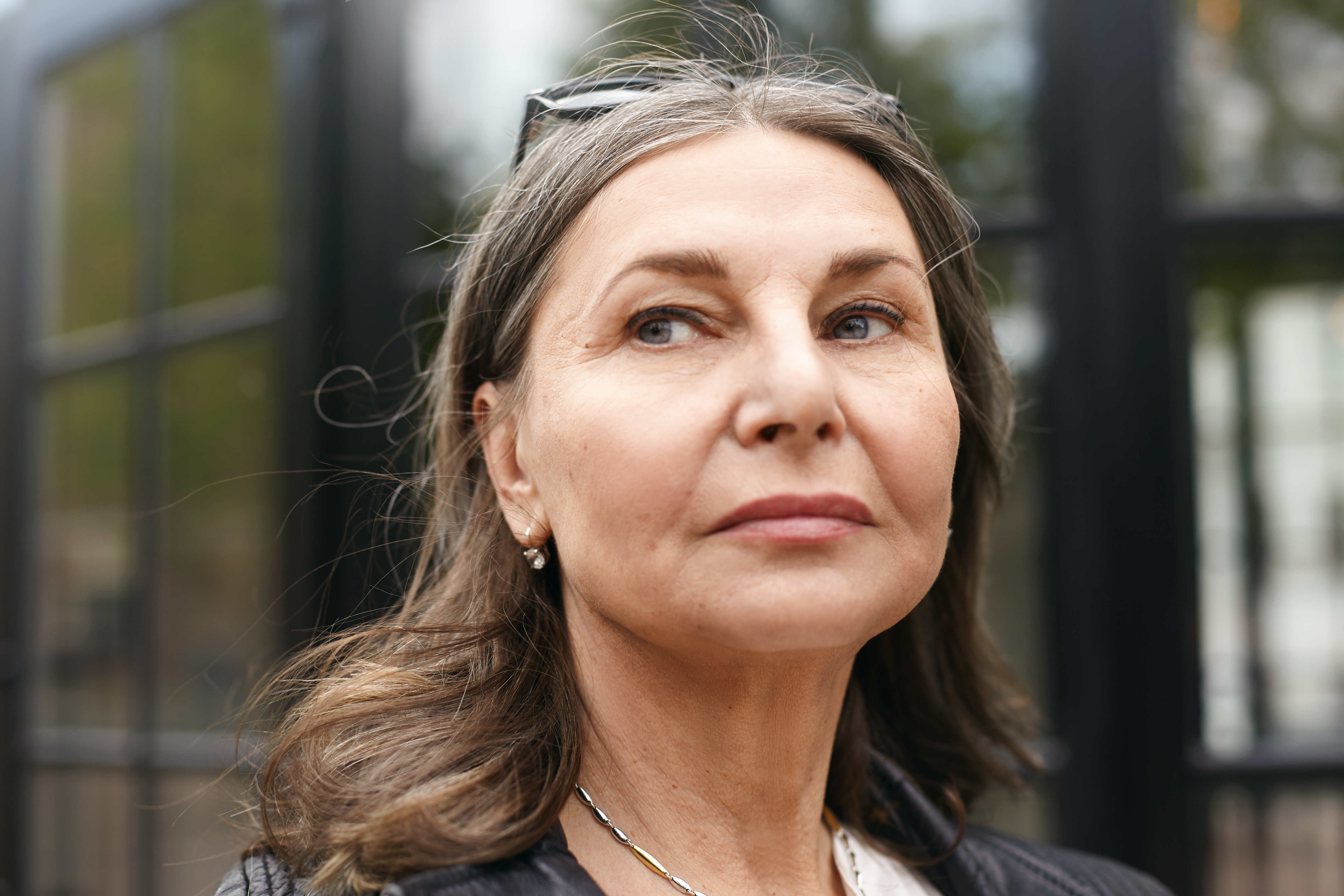 Close up portrait of attractive Caucasian female pensioner with loose gray hair and wrinkles having serious facial expression, looking away, posing outdoors with modern building in blurred background
